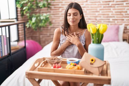 Photo for Brunette young woman eating breakfast sitting on the bed smiling with hands on chest with closed eyes and grateful gesture on face. health concept. - Royalty Free Image