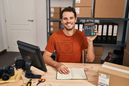 Foto de Joven hombre hispano que trabaja en el comercio electrónico de las pequeñas empresas sosteniendo la calculadora con un aspecto positivo y feliz de pie y sonriendo con una sonrisa confiada mostrando los dientes - Imagen libre de derechos