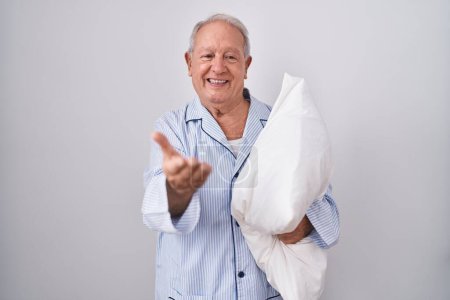 Foto de Hombre mayor con pelo gris usando pijama abrazando almohada sonriente alegre ofreciendo mano de palma dando asistencia y aceptación. - Imagen libre de derechos