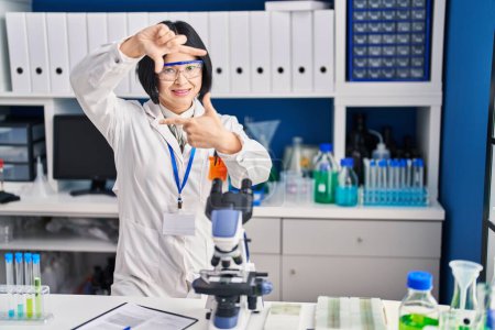 Téléchargez les photos : Jeune femme asiatique travaillant au laboratoire scientifique souriant faisant cadre avec les mains et les doigts avec le visage heureux. concept de créativité et photographie. - en image libre de droit