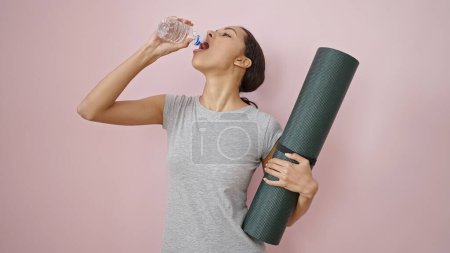 Photo for Young beautiful hispanic woman wearing sportswear holding yoga mat drinking water over isolated pink background - Royalty Free Image