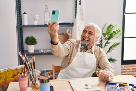 Téléchargez les photos : Moyen Age homme aux cheveux gris artiste souriant confiant faire selfie par smartphone au studio d'art - en image libre de droit