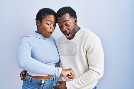 Téléchargez les photos : Jeune couple afro-américain debout sur fond bleu regardant l'heure de la montre inquiet, peur de se faire tard - en image libre de droit
