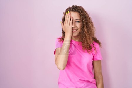 Foto de Young caucasian woman standing over pink background covering one eye with hand, confident smile on face and surprise emotion. - Imagen libre de derechos