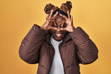 Photo for African woman with braided hair standing over yellow background doing ok gesture like binoculars sticking tongue out, eyes looking through fingers. crazy expression. - Royalty Free Image