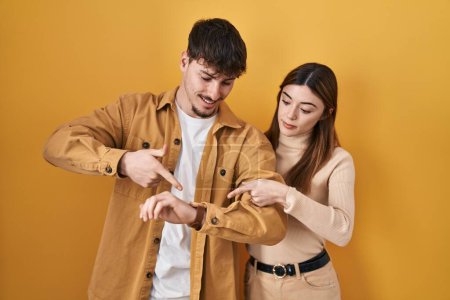 Téléchargez les photos : Jeune couple hispanique debout sur fond jaune à la hâte pointant pour regarder le temps, l'impatience, bouleversé et en colère pour retard de délai - en image libre de droit