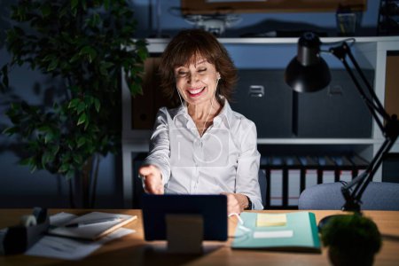 Téléchargez les photos : Femme d'âge moyen travaillant au bureau la nuit souriant amical offrant poignée de main comme salutation et accueil. entreprise prospère. - en image libre de droit