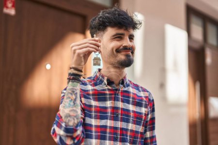 Téléchargez les photos : Jeune homme hispanique souriant confiant tenant la clé de la nouvelle maison à la rue - en image libre de droit