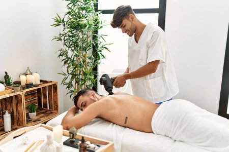 Photo for Two hispanic men physiotherapist and patient massaging back using percussion gun at beauty center - Royalty Free Image
