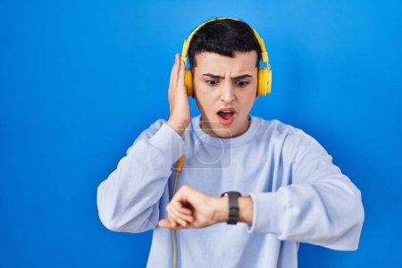 Foto de Persona no binaria escuchando música usando auriculares mirando la hora del reloj preocupada, temerosa de llegar tarde - Imagen libre de derechos