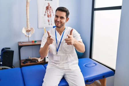 Photo for Young hispanic man with beard working at pain recovery clinic pointing fingers to camera with happy and funny face. good energy and vibes. - Royalty Free Image