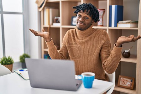 Photo for Young african man with dreadlocks working using computer laptop clueless and confused expression with arms and hands raised. doubt concept. - Royalty Free Image