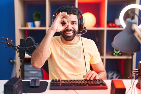 Photo for Hispanic man with beard playing video games with headphones doing ok gesture with hand smiling, eye looking through fingers with happy face. - Royalty Free Image