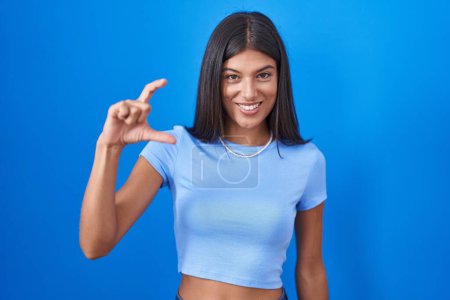 Téléchargez les photos : Brunette jeune femme debout sur fond bleu geste souriant et confiant avec la main faisant signe de petite taille avec les doigts regardant et la caméra. concept de mesure. - en image libre de droit