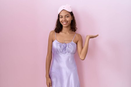 Photo for Young hispanic woman wearing sleep mask and nightgown smiling cheerful presenting and pointing with palm of hand looking at the camera. - Royalty Free Image