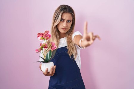 Foto de Mujer rubia joven con delantal de jardinero sosteniendo la planta señalando con el dedo hacia arriba y la expresión enojada, mostrando ningún gesto - Imagen libre de derechos