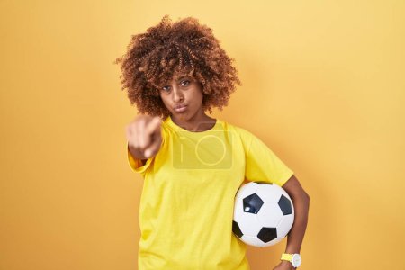 Photo for Young hispanic woman with curly hair holding football ball pointing with finger to the camera and to you, confident gesture looking serious - Royalty Free Image