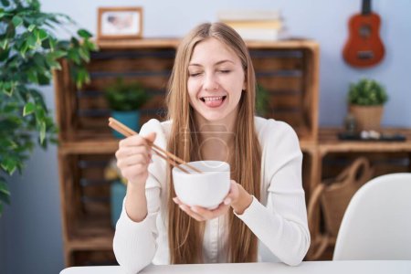 Foto de Joven mujer caucásica comiendo comida asiática usando palillos sacando la lengua feliz con expresión divertida. - Imagen libre de derechos
