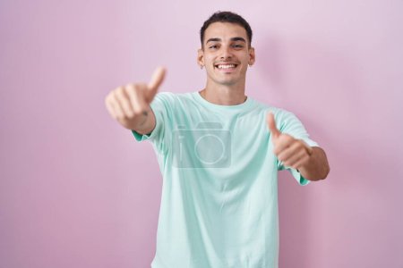 Foto de Hombre hispano guapo de pie sobre fondo rosa aprobando hacer gesto positivo con la mano, pulgares hacia arriba sonriendo y feliz por el éxito. gesto ganador. - Imagen libre de derechos