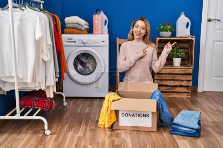 Photo for Hispanic woman putting clothes in donation box smiling and looking at the camera pointing with two hands and fingers to the side. - Royalty Free Image