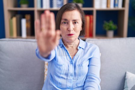 Photo for Middle age hispanic woman sitting on the sofa at home doing stop sing with palm of the hand. warning expression with negative and serious gesture on the face. - Royalty Free Image