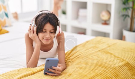 Foto de Adorable chica hispana escuchando música tumbada en la cama en el dormitorio - Imagen libre de derechos