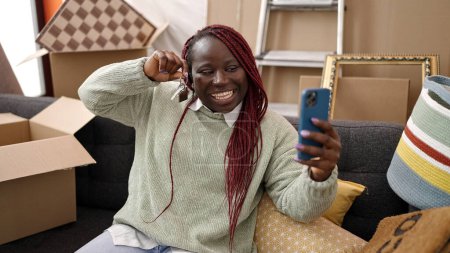 Téléchargez les photos : Femme africaine avec des cheveux tressés faire appel vidéo assis sur le canapé à la nouvelle maison - en image libre de droit