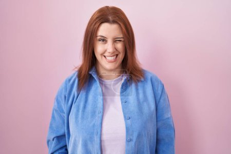 Foto de Mujer hispana joven con el pelo rojo de pie sobre fondo rosa guiño mirando a la cámara con expresión sexy, cara alegre y feliz. - Imagen libre de derechos