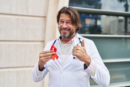 Téléchargez les photos : Beau médecin d'âge moyen homme tenant soutien ruban rouge sourire heureux et positif, pouce levé faire excellent et signe d'approbation - en image libre de droit