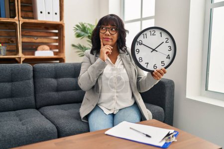 Photo for Hispanic woman working at therapy office holding clock serious face thinking about question with hand on chin, thoughtful about confusing idea - Royalty Free Image