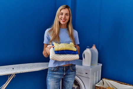 Photo for Young blonde woman smiling confident holding folded clothes at laundry room - Royalty Free Image