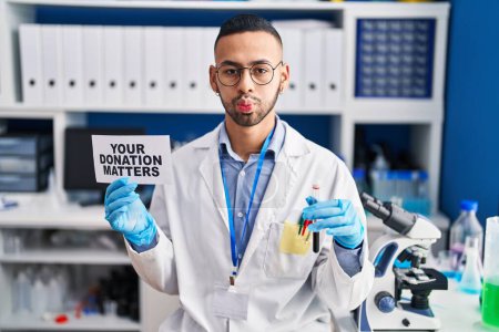 Photo for Young hispanic man working at scientist laboratory holding your donation matters holding blood sample puffing cheeks with funny face. mouth inflated with air, catching air. - Royalty Free Image