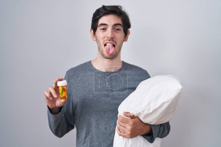 Foto de Joven hombre hispano usando pijama abrazando almohada sosteniendo píldoras sacando la lengua feliz con expresión divertida. - Imagen libre de derechos