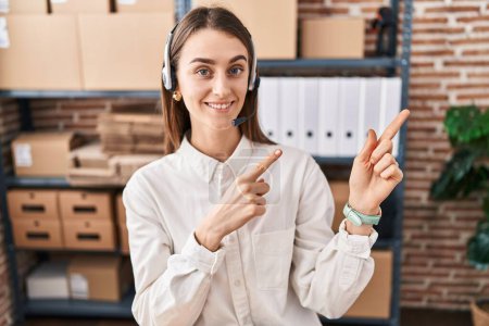 Photo for Young caucasian woman working at small business ecommerce wearing headset smiling and looking at the camera pointing with two hands and fingers to the side. - Royalty Free Image