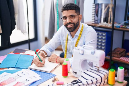 Téléchargez les photos : Jeune homme arabe tailleur souriant dessin confiant sur ordinateur portable au magasin de tailleur - en image libre de droit