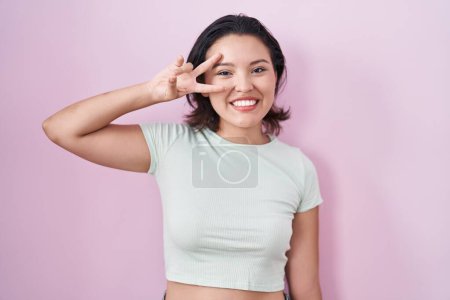 Téléchargez les photos : Jeune femme hispanique debout sur fond rose faisant symbole de paix avec les doigts sur le visage, souriant joyeux montrant la victoire - en image libre de droit