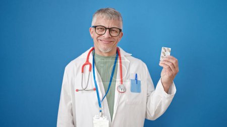 Téléchargez les photos : Moyen âge homme aux cheveux gris médecin tenant des pilules sur fond bleu isolé - en image libre de droit
