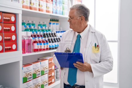 Téléchargez les photos : Homme moyen aux cheveux gris pharmacien écrivant sur un document à la pharmacie - en image libre de droit