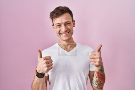 Téléchargez les photos : Homme caucasien debout sur fond rose signe de succès faisant geste positif avec la main, pouces levés souriant et heureux. expression joyeuse et geste gagnant. - en image libre de droit