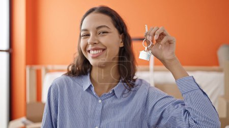 Foto de Joven hermosa mujer hispana sonriendo confiada sosteniendo nuevas llaves de la casa en un nuevo hogar - Imagen libre de derechos