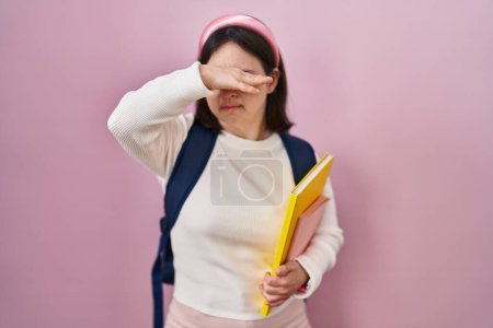 Photo for Woman with down syndrome wearing student backpack and holding books covering eyes with hand, looking serious and sad. sightless, hiding and rejection concept - Royalty Free Image