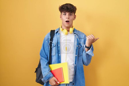 Foto de Adolescente hispana con mochila de estudiante y libros sorprendidos apuntando con el dedo de la mano hacia un lado, boca abierta expresión asombrada. - Imagen libre de derechos