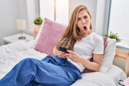 Téléchargez les photos : Jeune femme blonde utilisant smartphone sur le lit dans le visage de choc, l'air sceptique et sarcastique, surpris avec la bouche ouverte - en image libre de droit