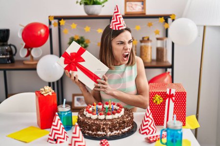 Foto de Mujer hispana celebrando cumpleaños con pastel sosteniendo regalo enojado y loco gritando frustrado y furioso, gritando con ira. rabia y concepto agresivo. - Imagen libre de derechos
