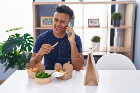 Téléchargez les photos : Jeune homme latino parlant sur smartphone manger emporter de la nourriture à la maison - en image libre de droit