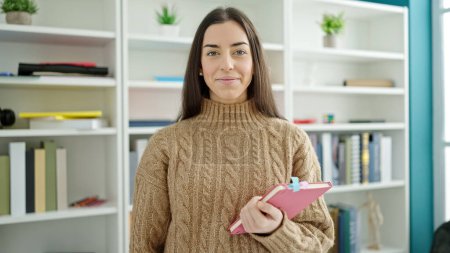 Photo for Young beautiful hispanic woman student smiling confident holding book at university classroom - Royalty Free Image
