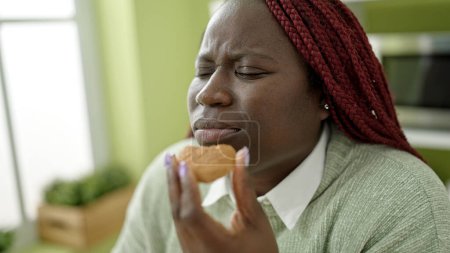 Téléchargez les photos : Femme africaine avec des cheveux tressés manger cookie avec les yeux fermés à la salle à manger - en image libre de droit