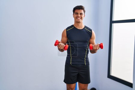 Photo for Young hispanic man smiling confident using dumbbells training at sport center - Royalty Free Image