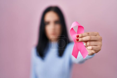 Foto de Mujer hispana sosteniendo actitud de pensamiento de cinta de cáncer rosa y expresión sobria con aspecto de confianza en sí misma - Imagen libre de derechos