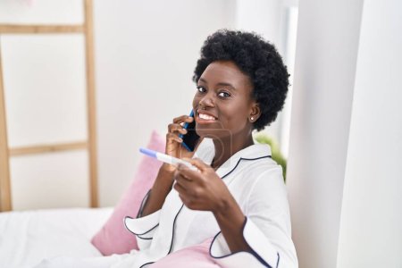 Photo for Young african american woman talking on the smartphone holding pregnancy test at bedroom - Royalty Free Image